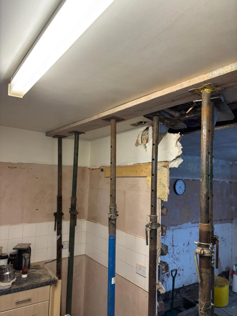 Adjustable steel acrow props support a wooden beam against a ceiling during a kitchen wall removal and home renovation.