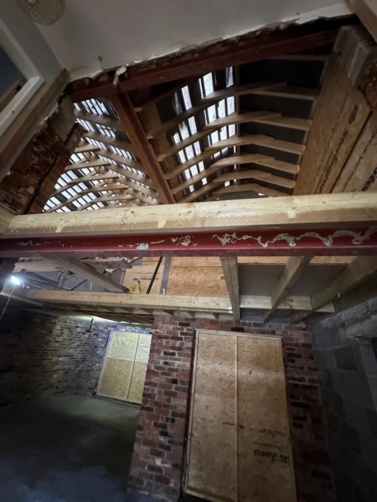 Internal view of a house renovation featuring exposed wooden roof rafters, steel beams, and brick walls.