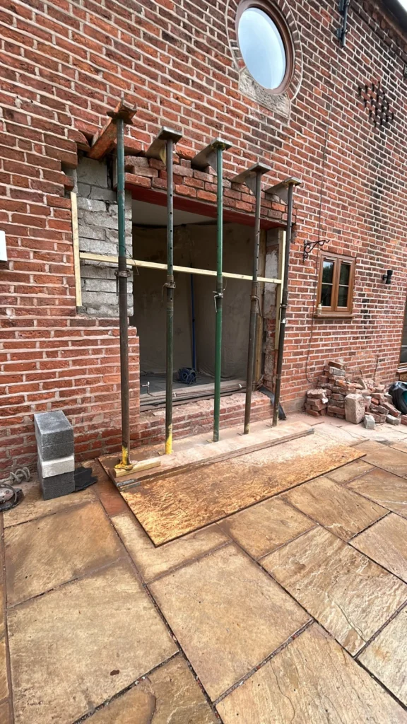 Five steel acrow props supporting a brick wall during a door installation on a stone patio.