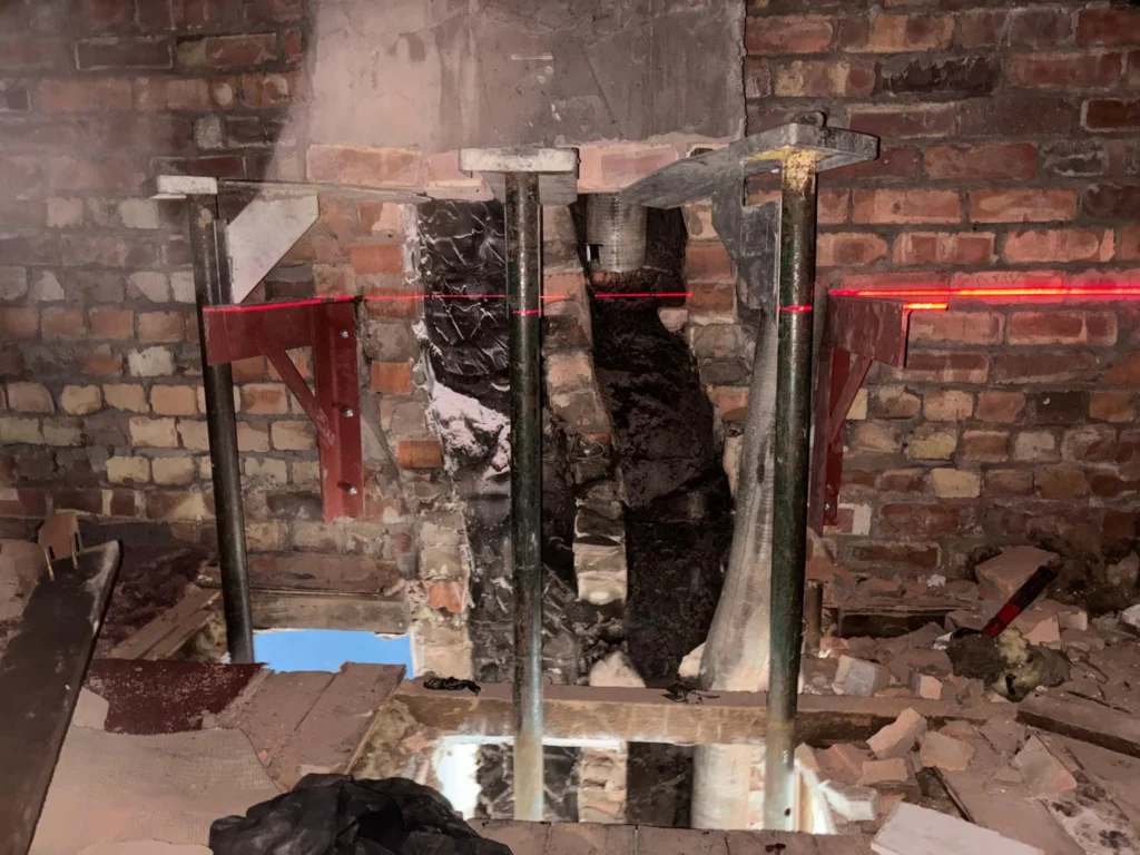 Steel acrow props and red brackets supporting a damaged brick wall during structural construction work.